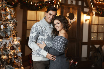 Beautiful couple of lovers in Scandinavian sweaters at New year interior. Sexy model girl and handsome man hugging together. A lovable young woman spending Christmas time with her husband.