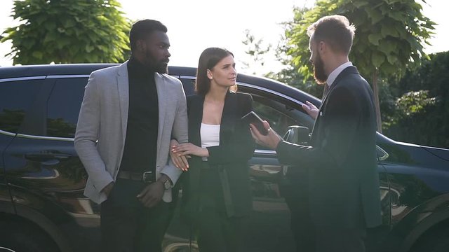 Salesman presenting a new car to young couple in outdoors. Successful car dealer tells the owners about the benefits of their car at parking near car dealership