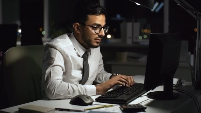 Medium shot of frustrated Arab businessman in glasses working overtime in dark office. He is typing on computer, then turning towards camera and ranting about something
