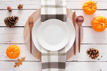 Fall harvest or thanksgiving dinner table setting with plate, flatware, check print napkin, pumpkins and decor. Above view on a white wood background.