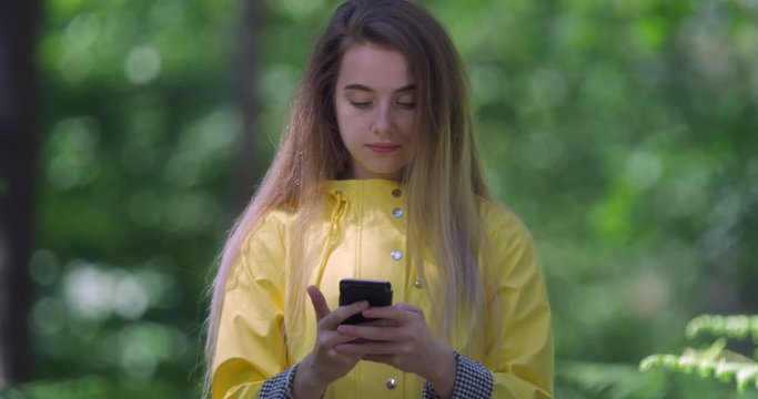  Young Smiling Woman with Mobile phone talking in a Forest. Girl in a Yellow Parka in the Woods. Pretty Hiker with chatting on Cell  within tree foliage at a Green Park with Natural Sun Light..
