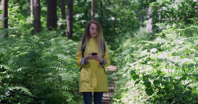 Young Woman with Mobile Phone in a Forest. Girl in a Yellow Coat in the Woods. Pretty Hiker using sat nav on Cell within tree foliage at a Green Park with Natural Sun Light