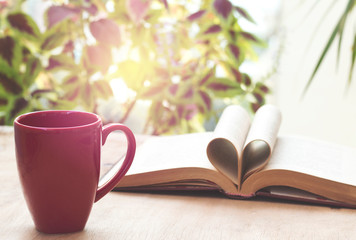 cup and open book on wooden table in front of the window