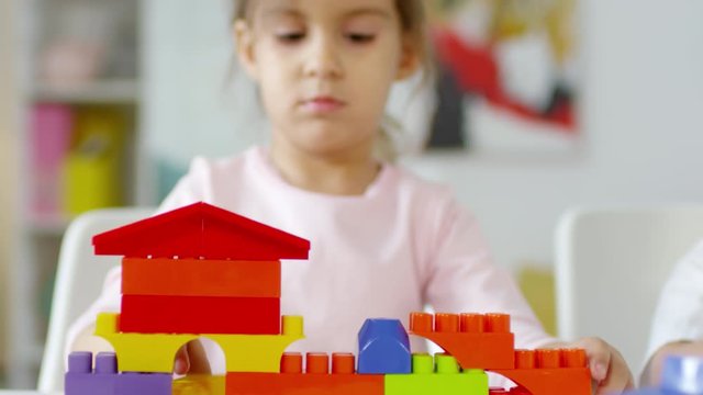 Rack focused shot of pretty little girl building toy house with colorful plastic bricks and smiling while playing with construction toy at table