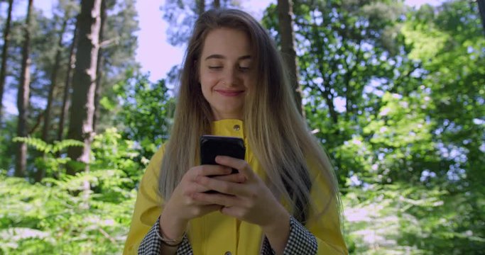 Young Smiling Woman with Mobile Phone in a Forest. Girl in a Yellow Rain Coat in the Woods. Pretty, blonde Student Girl using a Cell within tree foliage in a Summer Green Park with Natural Sun Light.