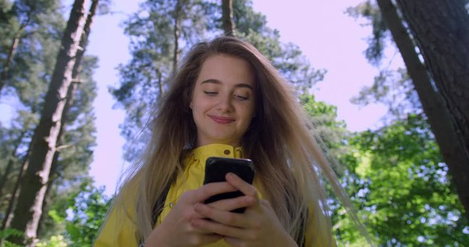 Young Smiling Woman with Mobile Phone in British Forest. Girl in Woodland Glade wearing a Yellow Rain Coat. Pretty, blonde Student Girl Swiping her Cell within Tree foliage in a Green Natural Park 