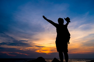 Fototapeta premium Traveler backpacker of freedom style Concept: Silhouette of Asian Young woman relaxing standing on rock seabeach, Strong confidence open her arms and watching sunset scene in summer sunset sky outdoor