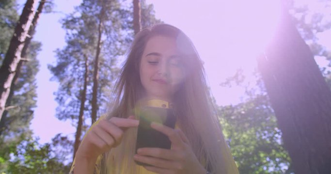  Young Smiling Woman swiping a Mobile Phone in the Forest. Girl in Woodland Glade wearing a Yellow Rain Coat. Happy, blonde Student Girl with Cell Emoji within Tree foliage at a Green Natural Park