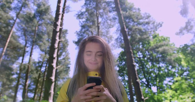 Young Smiling Woman swiping a Mobile Phone in the Forest. Girl in Woodland Glade wearing a Yellow Rain Coat. Happy, blonde Student Girl with Cell Emoji within Trees at a Green Natural Park