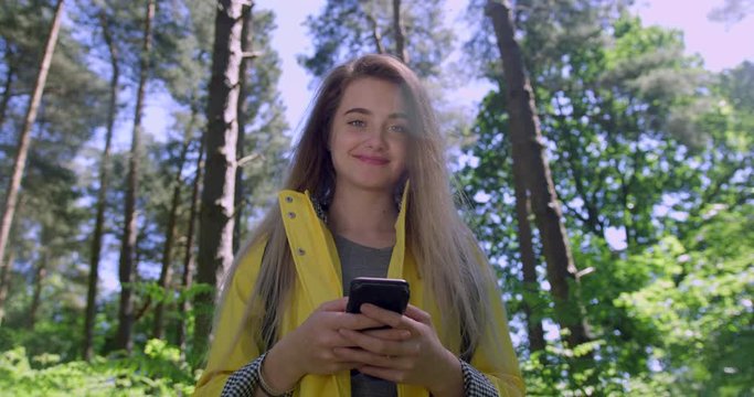 Young Woman texting with Mobile Phone in the Forest. Girl in Wood land Glade wearing a Yellow Rain Coat. Happy, blonde Student Girl with Cell Emoji within Trees at a Green Natural Park