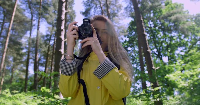 Young Woman with Camera in the Forest. Girl taking Photographs in a Woodland Glade wearing a Yellow Rain Coat. Happy, blonde Student Girl within Trees at a Green Natural Park with Sun Light Flare...