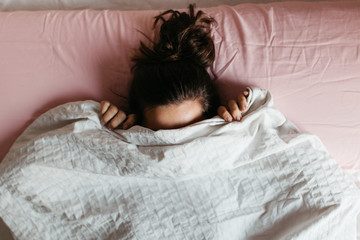 Playful young woman hiding face under blanket while lying in cozy bed on white pillow, pretty...