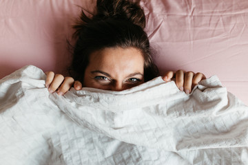 Playful young woman with beautiful eyes hiding face under blanket while lying in cozy bed, pretty...