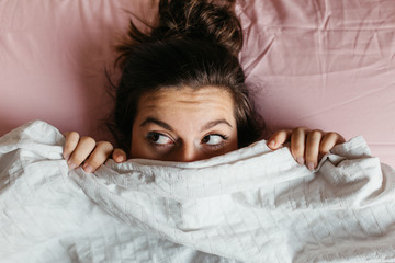 Scared and surprised young woman with open eyes hiding face under blanket, pretty frightened and...