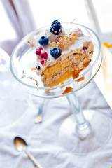 A piece of cake from biscuit and white cream with forest berries on top served on glass high stand on white cloth
