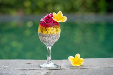 Chia seeds pudding with red dragon fruit, passion fruit, mango and avocado in a glass for breakfast on the background of the swimming pool water, closeup. The concept of healthy eating.