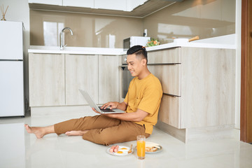 Fototapeta na wymiar Young business man is smiling, eating breakfast and working on laptop in bright kitchen.