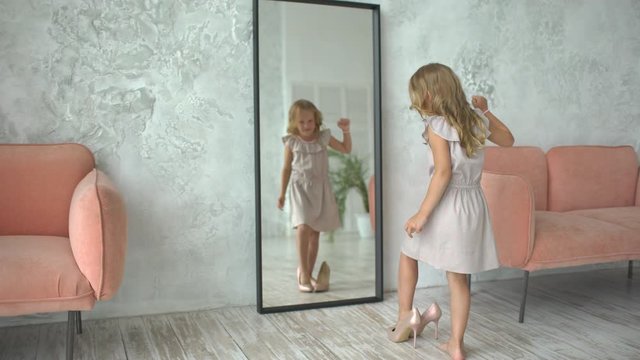 Cute pretty little girl in dress child fashionista in high heels with big mother's shoes looking in mirror at home. child in women's adult mom's shoes. Glamour Fashion Beauty Girl Model small, 4 K