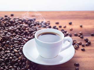 Natural roasted coffee beans pilled on brown table, close up view. White coffee cup with black espresso on wooden table. Texture of wooden desk. Selective soft focus. Blurred background
