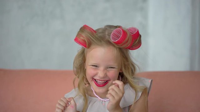 Portrait of little funny girl in hair curlers applying lipstick smiling and laughing. child with lipstick, playing with mothers makeup. Face of blonde little fashionista having fun, painting lips, 4 K