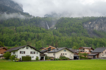 Fototapeta na wymiar Houses beside the misty mountain full of fresh green trees with waterfall in rural area of Switzerland for background and copy space