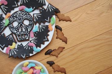 Halloween cookies in shape of skull and bat with colorful worm candies on a plate on wooden background. Halloween sweet food