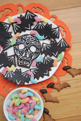 Halloween cookies in shape of skull and bat with colorful worm candies on a plate on wooden background. Halloween sweet food