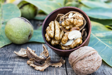 Walnut kernels lie in a bowl next to nuts in green shells and green leaves on a rustic old wooden table.