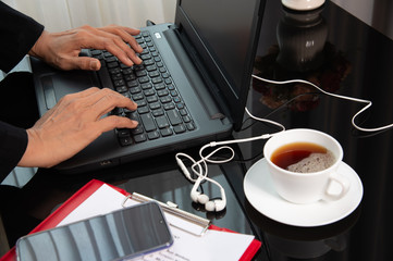 Working woman in black suit typing computer surrounds with smartphone, red note, white earphone and a cup of coffee on table in the office.