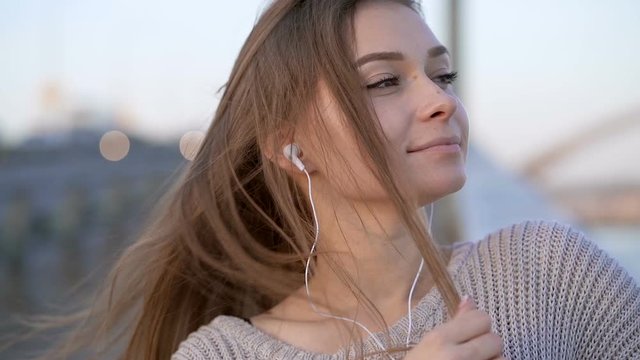 Close up portrait of young beatiful woman outdoors. Standing at the street. Listening to music in the hradphones and relaxing. Handheld shot. Slowmotion.