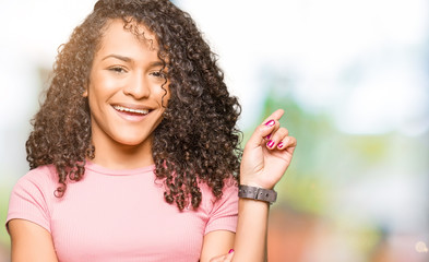 Young beautiful woman with curly hair wearing pink t-shirt with a big smile on face, pointing with hand and finger to the side looking at the camera.