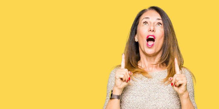 Beautiful middle age woman wearing fashion sweater amazed and surprised looking up and pointing with fingers and raised arms.