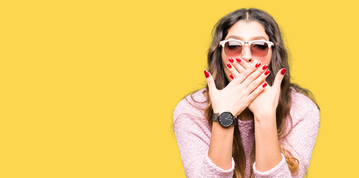 Young beautiful woman wearing sunglasses and pink sweater shocked covering mouth with hands for mistake. Secret concept.