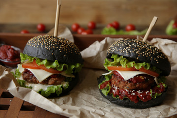 Two black sandwich, hamburger or burger with beef or chicken, tomatoes, onions, cheese, lettuce and a sesame bun on a paper on a wooden rustic board. Fast food concept