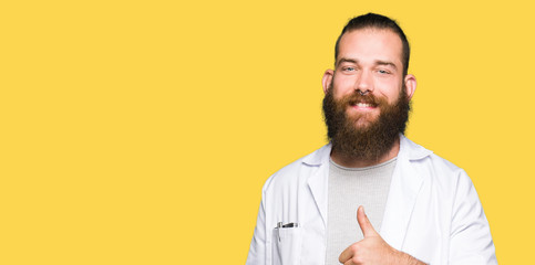 Young blond scientist man wearing white coat doing happy thumbs up gesture with hand. Approving expression looking at the camera with showing success.