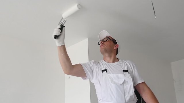 Painter worker with roller painting ceiling surface into white