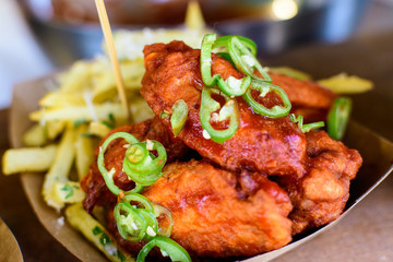 Hot Buffalo chicken wings with French fried potatoes and  chopped jalapenos peppers in brown paper, displayed for sale at a street food market in direct sunlight, selective focus