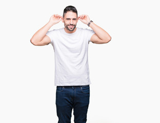 Fototapeta premium Handsome man wearing white t-shirt over white isolated background Smiling pulling ears with fingers, funny gesture. Audition problem