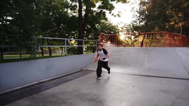 Sportive man practicing parkour in the open skate park - sliding from the ramp, crossing the barries amd making somersault in slow motion while holding a red coloured smoke bomb in a hand. Summer day