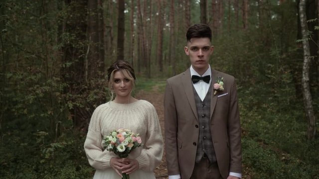 A beautiful girl and a guy in wedding dresses stand and look into the camera. Average plan. Wedding couple without emotion looking at the camera. Serious emotions
