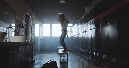 Camera follows happy athletic European woman entering dark gym locker room dancing, feeling excited after good workout.
