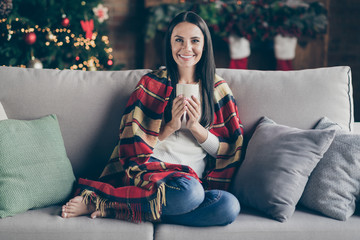 Full size photo of positive cheerful girl sit divan barefoot over with checkered blanket enjoy christmas time holidays hold mug with hot eggnog in house with x-mas decoration lights indoors