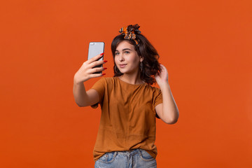 Pretty brunette woman in a t-shirt and beautiful headband making selfie with spartphone standing isolated over orange background. Mobile photography