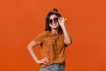 Pretty brunette woman in a t-shirt and beautiful headband expressing emotion of surprise holding sunglasses standing isolated over orange background.