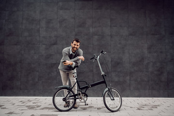 Fototapeta na wymiar Full length of smiling handsome caucasian fashionable man leaning on his bicycle and holding smart phone and glasses. In background is gray wall.