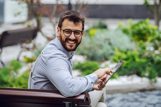 Smiling Attractive Caucasian Businessman With Eyeglasses Sitting On Bench In Park And Using Tablet While Looking Over Shoulder.