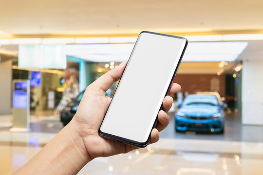 Mockup image of hand holding blank screen mobile smart phone with blurred background of new cars display in showroom, buying new car. Mock up smartphone for your advertisement.