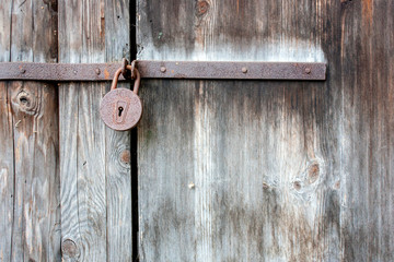 A rusty lock hangs on closed doors from old planks. Gray door boards and rusty steel hinges closeup.