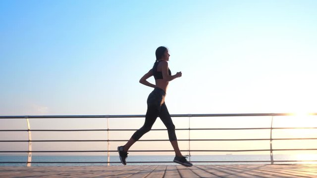 Athletic young girl jogging in the morning by sea wooden embankment. Silhouette of girl in sports costume. Beautiful sunlight. Healthy lifestyle concept