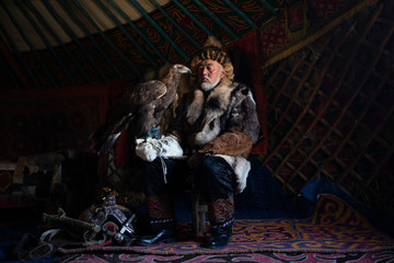 Portrait of an old kazakh eagle hunter with his majestic golden eagle indoors in dark traditional...